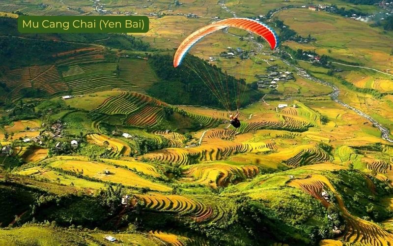 Paraglider soaring over the terraced rice fields of Mu Cang Chai in Yen Bai, Vietnam, with an orange-and-white canopy above layered golden-green fields, winding roads, scattered houses, and mountain slopes, labeled “Mu Cang Chai (Yen Bai)” in the top left corner.