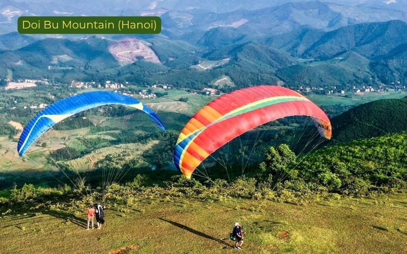 Two colorful paragliders flying near Doi Bu Mountain outside Hanoi, Vietnam, above rolling green hills and a valley landscape, with red-and-blue canopies visible in the foreground and the label “Doi Bu Mountain (Hanoi)” in the top left corner.