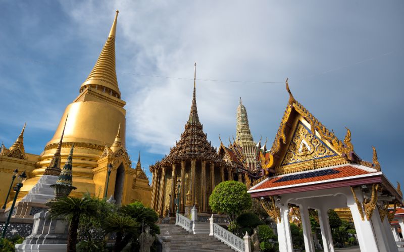 A wide panoramic view under a dramatic sky, capturing the immense golden Phra Siratana Chedi, the detailed library building (Phra Mondop), and the tiled roofs of the complex from a surrounding courtyard.