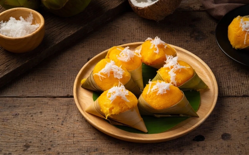 Cone-shaped Cambodian palm sugar desserts wrapped in banana leaf, topped with grated coconut, served on a wooden plate.
