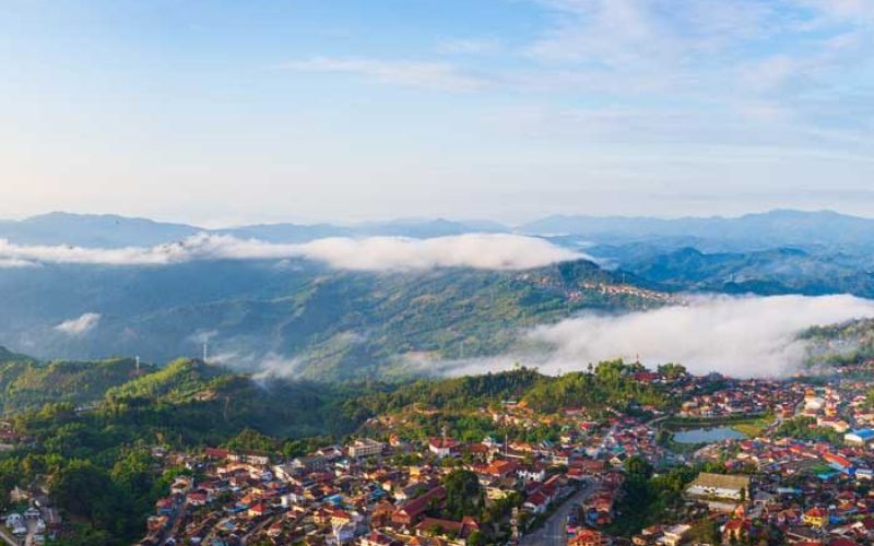 A scenic view of Phongsali, Laos, showing a small town nestled among green hills with morning mist and clouds hovering over the landscape.