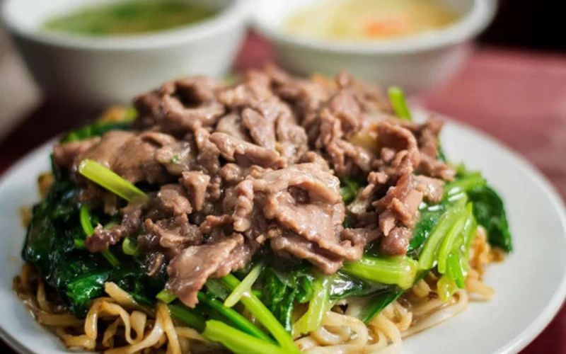 Plate of pho xao (stir-fried pho noodles) topped with beef and greens, served hot with dipping sauce on the side