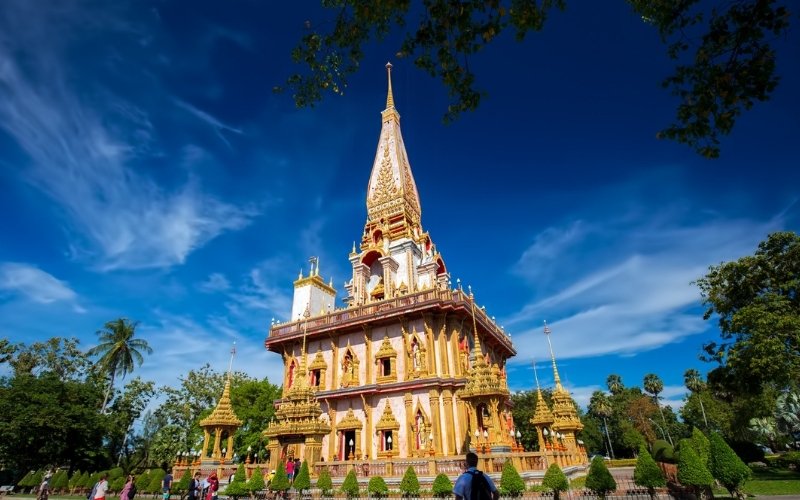 A low-angle shot of the majestic Grand Pagoda at Wat Chalong in Phuket, showcasing its intricate pink and gold architecture against a deep blue sky.