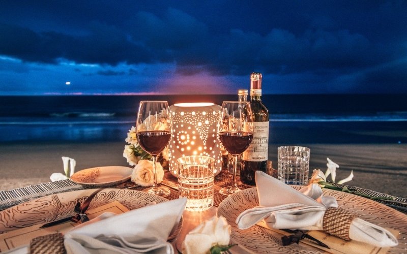 Elegant private dinner setup on a beach in Thailand with wine glasses, candles, and flowers, overlooking the ocean at sunset.