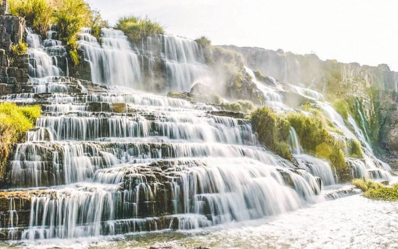 A wide-angle view of Pongour Waterfall in Lam Dong, Vietnam, showing its seven terraced rock layers cascading down into a rocky pool, framed by lush green forest under a cloudy highland sky.