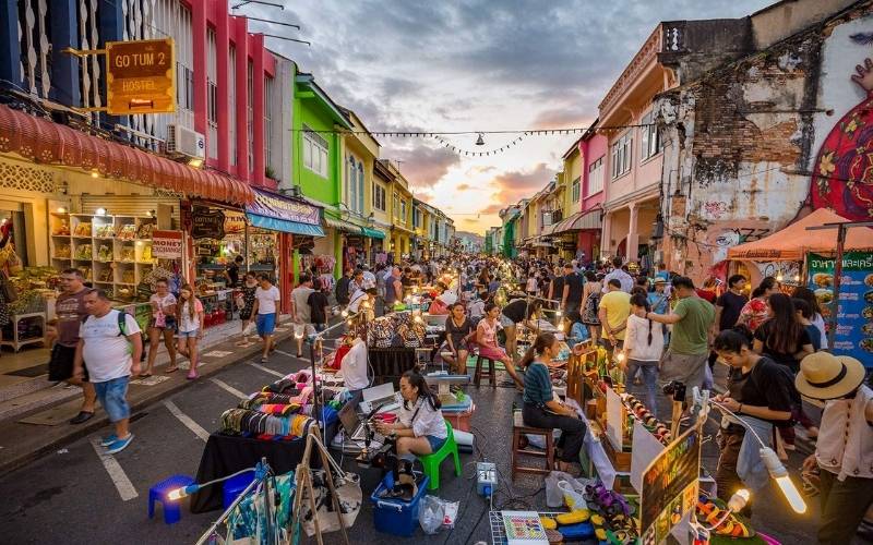 A bustling crowd fills the street at the Phuket Old Town Walking Street Weekend Market, surrounded by colorful Sino-Portuguese shophouses and vendors selling local goods at sunset.