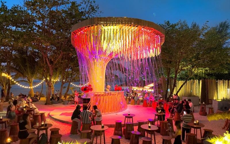 A dynamic night shot of a large crowd dancing at a beach party in Phuket, illuminated by vibrant neon stage lights with a fire dancer performing in the foreground.