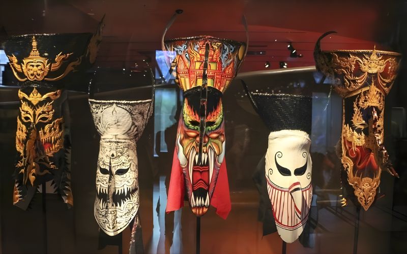 Display of traditional Phi Ta Khon masks from Thailand, featuring several tall ghost festival masks with long noses, horn-like tops, and intricate painted designs in black, red, white, and gold, exhibited indoors under museum lighting.