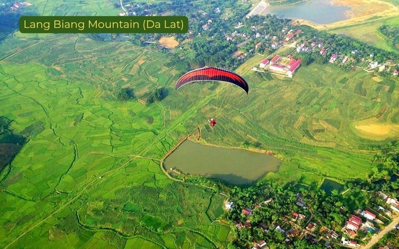 Aerial paragliding view over Lang Biang Mountain in Da Lat, Vietnam, showing a solo paraglider with a red-and-black canopy flying above patchwork green fields, a small lake, scattered houses, and a rural landscape, with the label “Lang Biang Mountain (Da Lat)” in the top left corner.