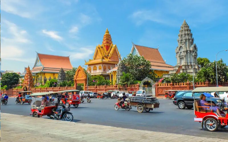 Royal Palace and Silver Pagoda complex in Phnom Penh, Cambodia, with tuk-tuks and motorbikes passing along the busy street in front of the ornate golden Khmer architecture.
