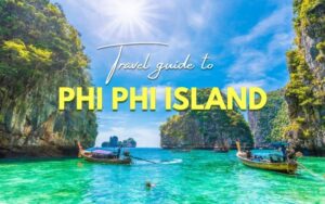 Phi Phi Island travel guide: A stunning tropical view of Phi Phi Island in Thailand, featuring turquoise water, traditional long-tail boats, and towering limestone cliffs under a bright blue sky with sunshine.