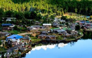 A peaceful lakeside village surrounded by lush green hills in Northern Thailand, with colorful rooftops reflecting on the water