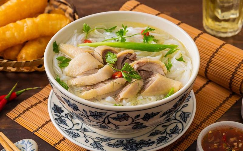 Bowl of pho ga (Vietnamese chicken noodle soup) served with fresh herbs, chili, and quẩy on a bamboo placemat