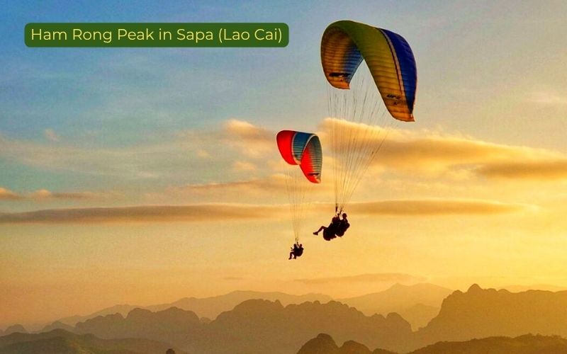 Sunset paragliding scene at Ham Rong Peak in Sapa, Lao Cai, Vietnam, showing two paragliders with colorful canopies flying above layered mountain silhouettes and a glowing evening sky, with the label “Ham Rong Peak in Sapa (Lao Cai)” in the top left corner.