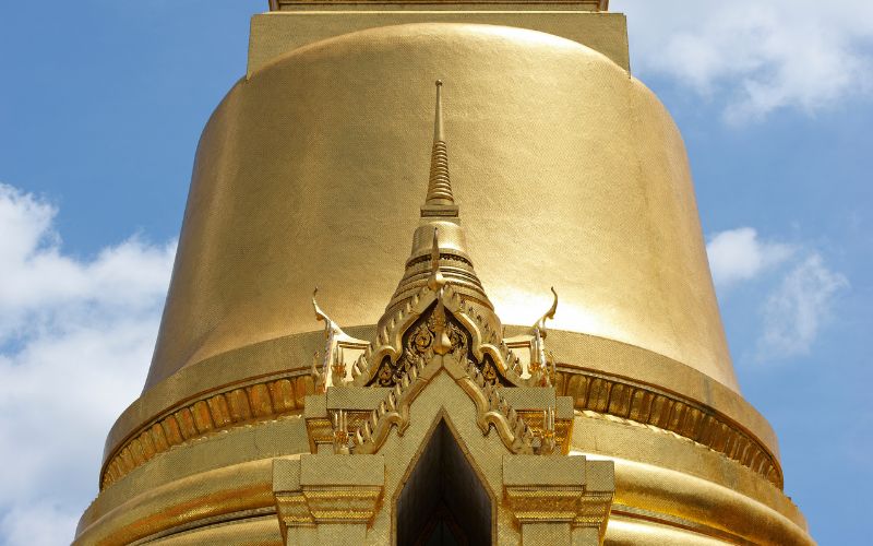 A tight close-up shot focused on the mid-section and towering golden spire of the Phra Siratana Chedi, emphasizing the texture of thousands of gold tiles and a complex ornamental niche against a blue sky.