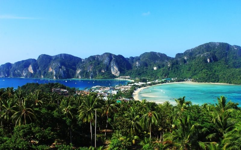 Panoramic view of Phi Phi Islands in Thailand, showing two curved bays with turquoise waters, white sand beaches, and lush green mountains under a clear blue sky.