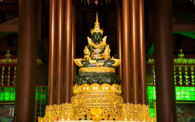 Inside a richly decorated temple hall with large dark wooden pillars, the venerated Emerald Buddha, carved from a single piece of jade and adorned in gold seasonal attire, sits enshrined on a high, tiered golden pedestal.