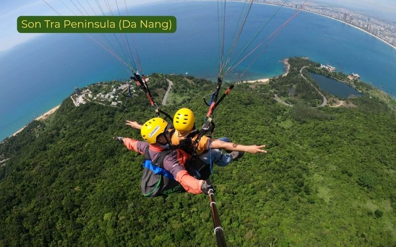 Tandem paragliding flight above Son Tra Peninsula in Da Nang, Vietnam, showing two riders wearing yellow helmets soaring over dense green forest, curved coastal roads, blue sea, and the Da Nang shoreline, with the label “Son Tra Peninsula (Da Nang)” in the top left corner.