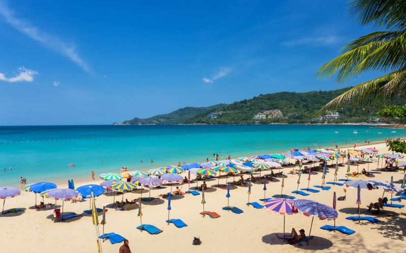 View of Patong Beach in Phuket, Thailand, with long sandy shoreline, and tourists relaxing by the sea.
