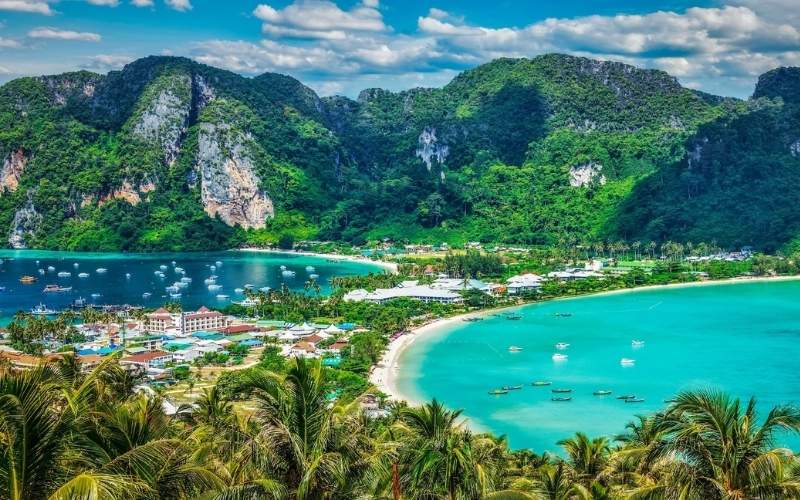 Scenic panoramic view of Phi Phi Island, Thailand, featuring lush green mountains, turquoise bays, white sandy beaches, and boats floating on the water under a bright blue sky.