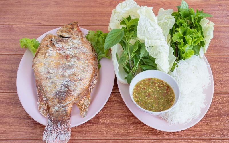 Thai pla pao served on two pink plates on a wooden table, with one whole salt-crusted grilled fish on the left and a side plate on the right containing fresh cabbage, lettuce, basil, rice vermicelli noodles, and a bowl of green spicy dipping sauce.