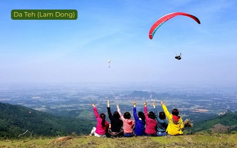 Scenic paragliding view in Da Teh, Lam Dong, Vietnam, showing a red paraglider high in a clear blue sky above a broad valley, while a group of people in colorful jackets sit on a grassy hillside watching and raising their hands, with the label “Da Teh (Lam Dong)” in the top left corner.