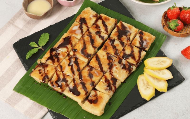 Sliced banana roti drizzled with condensed milk and chocolate sauce, served on a banana leaf with fresh strawberries and bananas.