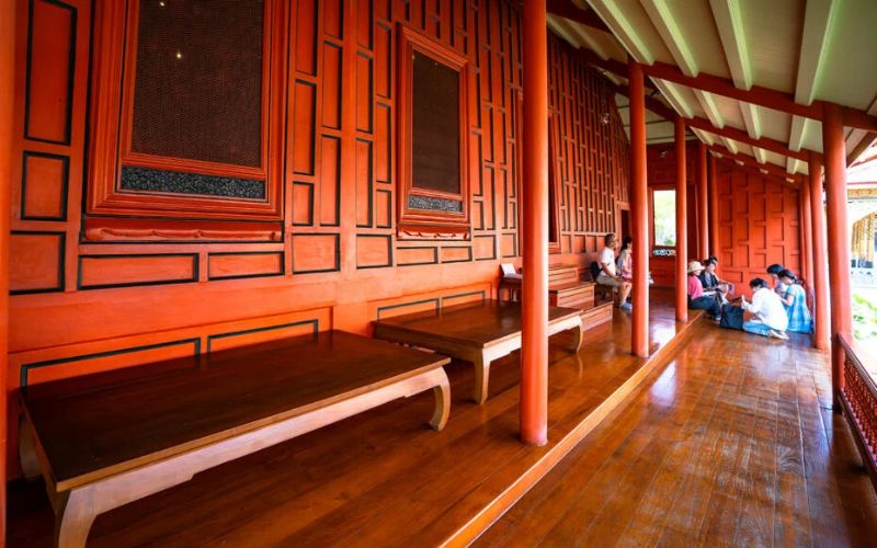 A long, shaded wooden veranda at the Red House (Tamnak Daeng) in the Bangkok National Museum, featuring vibrant reddish-orange paneled walls, polished floors, and visitors resting on the raised platform.