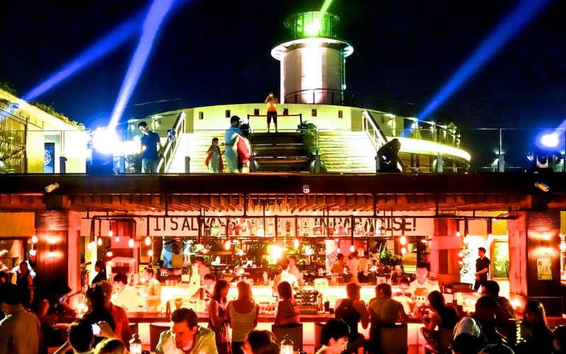 Tourists enjoying drinks at a lively rooftop bar in Nha Trang with neon lights and a view of the coastline