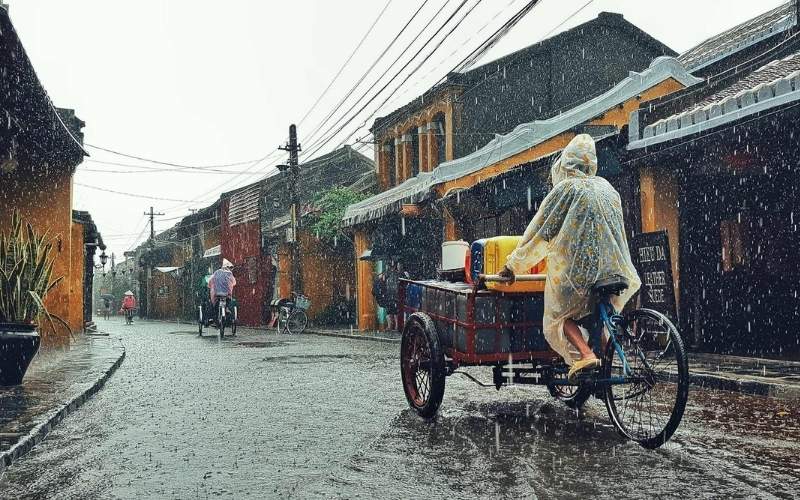 A person wearing a raincoat rides a bicycle cart through a wet street in Hoi An during heavy rain, surrounded by old yellow houses typical of southern Vietnam.