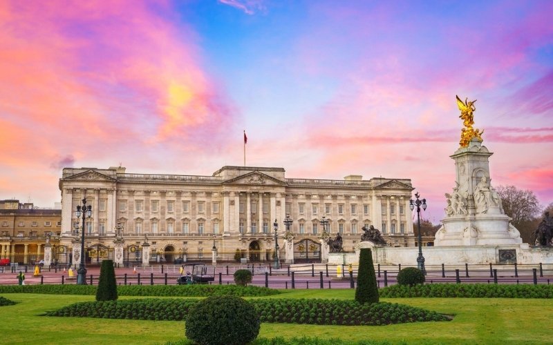 A grand palace building with elegant architecture, a central flag flying on top, and a golden statue monument in front, under a colorful sunset sky.