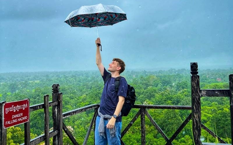 A traveler holding an umbrella stands on a wooden viewpoint overlooking lush green forests in Cambodia during a rainy day.