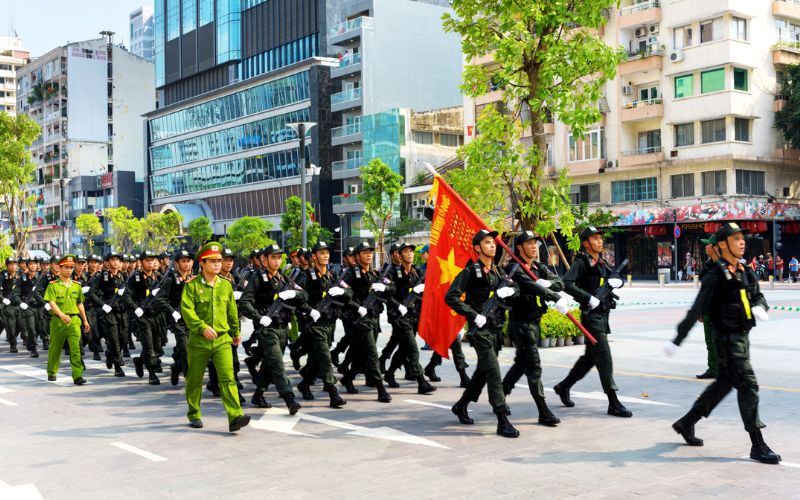 Reunification Day parade in Ho Chi Minh City, Vietnam, showing a formation of marching soldiers in black uniforms carrying rifles and the Vietnamese national flag, accompanied by officers in green uniforms, moving along a wide city street lined with trees, apartments, and modern buildings.