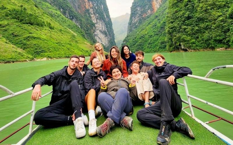 A group of happy travelers sitting together on a boat surrounded by lush green mountains and a peaceful river in northern Vietnam, smiling and making peace signs.