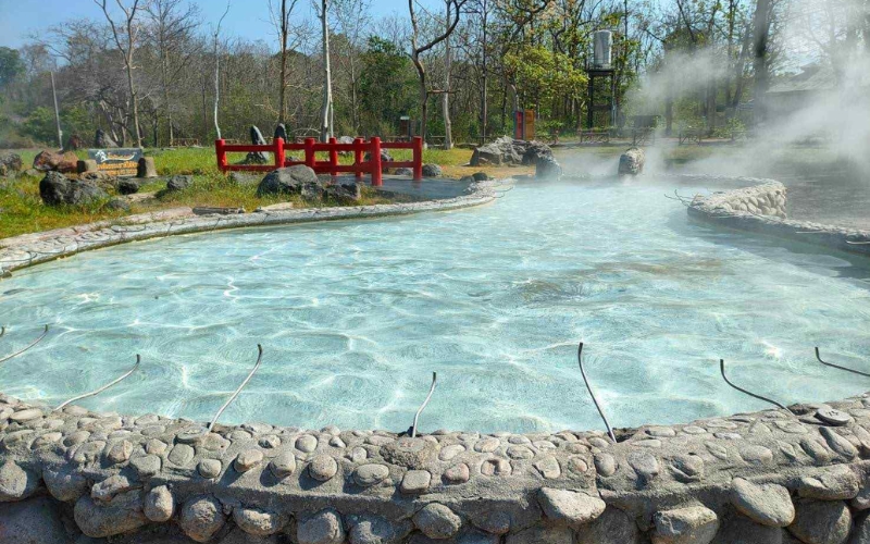 A tranquil outdoor scene showing a large, clear pool of steaming, light-blue water surrounded by a natural rock border and grass, indicating the location of the Sankhampaeng Hot Springs. Steam rises visibly over the water.