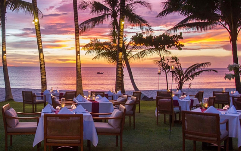 A beachfront restaurant in Phuket, with the sun setting over the Andaman Sea and casting golden reflections on the water.