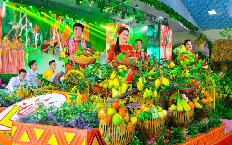 Southern Fruit Festival stage performance in Ho Chi Minh City, Vietnam, featuring performers in colorful traditional costumes carrying baskets of fruit, surrounded by decorative displays of mangoes and other tropical fruits, flowers, and bright green stage lighting.