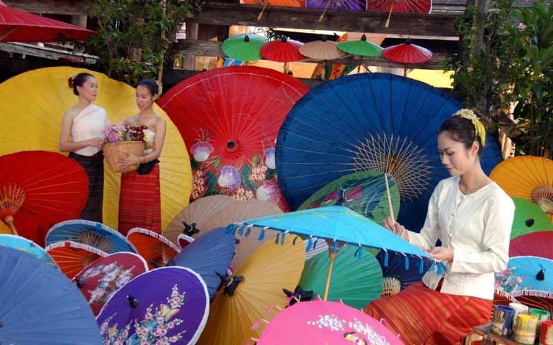 A beautiful, colorful display of numerous traditional Thai paper umbrellas (parasols) suspended overhead, creating a vibrant canopy of pink, orange, green, yellow, and blue at the Bo Sang and Sankhampaeng craft festival.