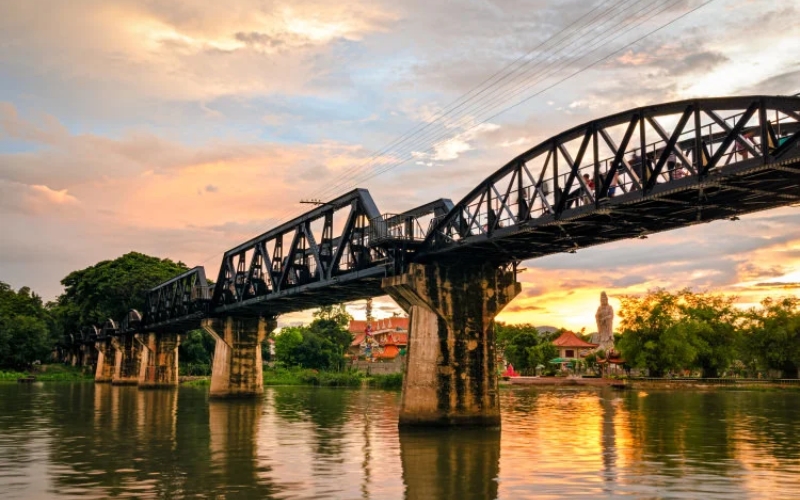 Golden sunset casting reflections over the River Kwai, with the iconic steel bridge silhouetted against the sky in Kanchanaburi, Thailand