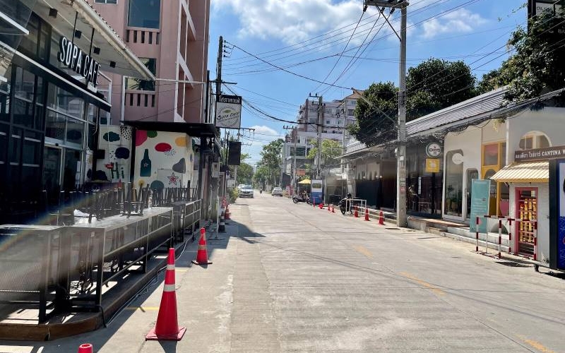 A daytime view of a quiet street on Nimmanhaemin Road in Chiang Mai, lined with modern cafes, shops, and low-rise commercial buildings under a bright blue sky.