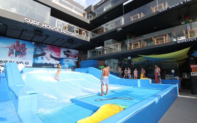A woman flowriding on an artificial wave machine at the Surf House Patong facility in Phuket, while people watch from the side near the modern, multi-story commercial building.