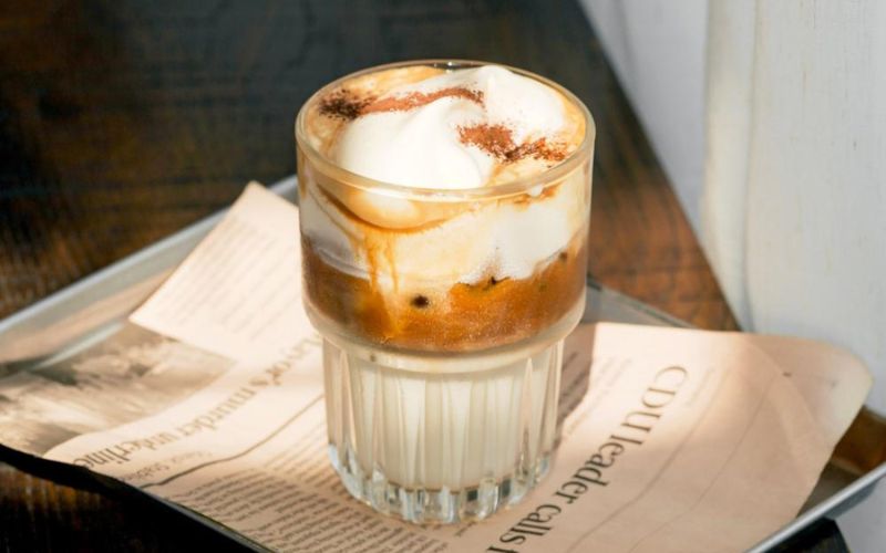 A glass of Vietnamese salted coffee with creamy foam and a sprinkle of cocoa, served on a newspaper-lined tray