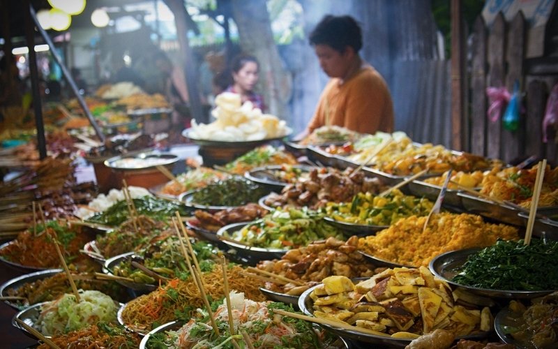 A close-up, dynamic shot of a Thai street food stall brimming with dozens of colorful dishes piled high on platters, including various stir-fries, noodles, fresh vegetables, and fried rice. Bamboo skewers and chopsticks stick out of the food. A vendor in an orange shirt is visible in the background, preparing food amidst a light haze of steam.