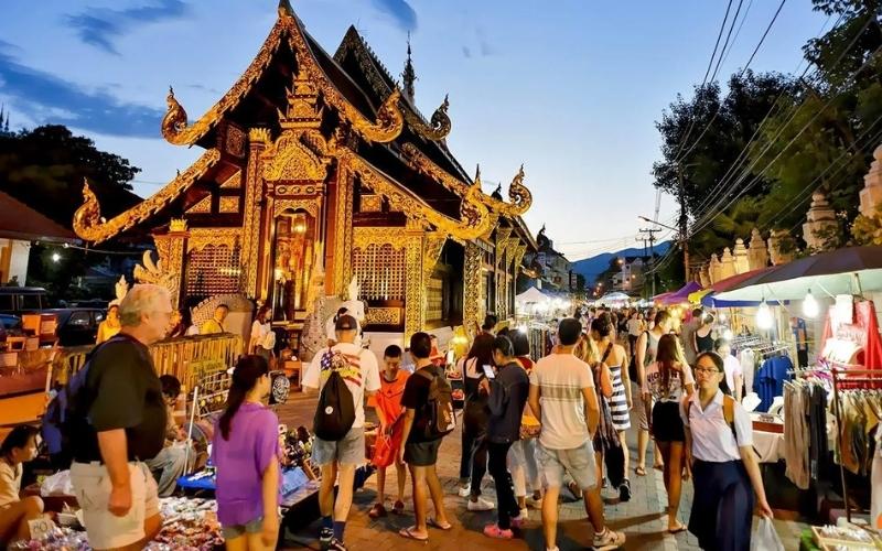 Crowds of tourists and locals browsing the Saturday Night Market on Wua Lai Street in Chiang Mai at twilight, walking past a beautifully illuminated golden Lanna-style temple.