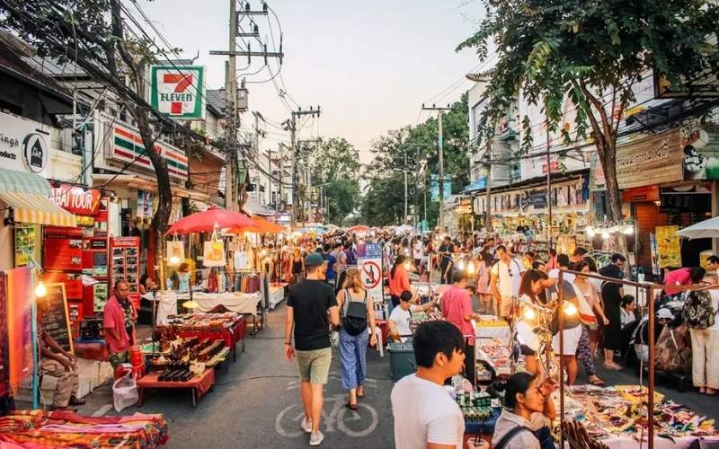 A wide shot of the busy Sunday Walking Street market in Chiang Mai, Thailand, featuring a crowd of pedestrians, a prominent 7-Eleven sign, and numerous stalls selling goods along the road.