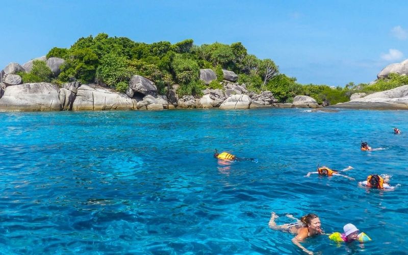 Snorkelers swimming in intensely clear, deep turquoise water near a small, rocky tropical island covered in lush green vegetation and large granite boulders under a bright blue sky.