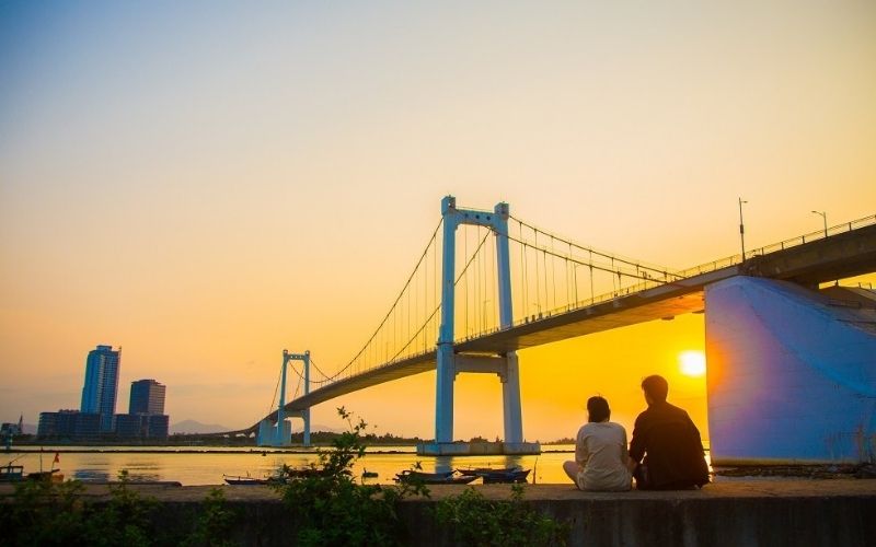 Romantic view of bridge, skyline, and glowing sun with a peaceful riverside ambiance of Han River, Da Nang