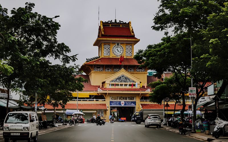 The front facade of Binh Tay Market, the largest commercial hub in Cho Lon, featuring a prominent central clock tower and a distinct yellow architectural style that blends Asian and classic French influences. The street in front is active with vehicles and lined with green trees.