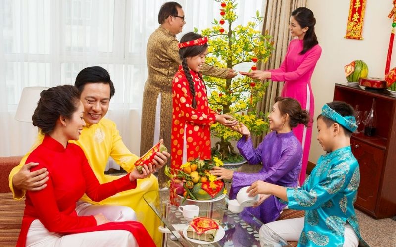 A Vietnamese family wearing colorful traditional áo dài, gathered in a living room during Lunar New Year, exchanging red envelopes and sharing joyful conversation around a decorated apricot blossom tree.