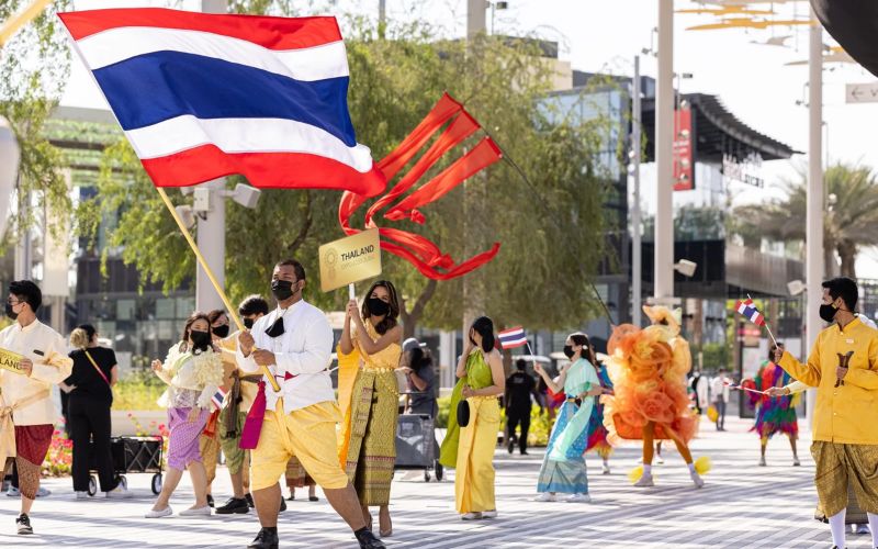 People celebrating Thailand National Day in traditional Thai costumes while carrying the Thai national flag during a cultural parade in a public square.